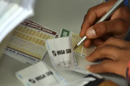 Pessoa preenche um volante de aposta da Mega-Sena com uma caneta, ao lado de outros bilhetes de loteria