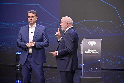 Lula gesticula durante debate presidencial, enquanto Flávio Bolsonaro observa ao seu lado, em estúdio da TV Bandeirantes