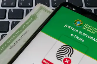 Celular com o aplicativo e-Título aberto sobre um título de eleitor impresso e um teclado de computador
