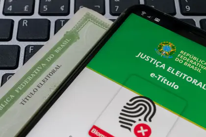 Celular com o aplicativo e-Título aberto sobre um título de eleitor impresso e um teclado de computador