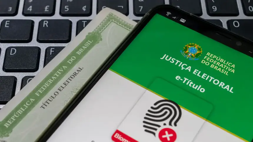 Celular com o aplicativo e-Título aberto sobre um título de eleitor impresso e um teclado de computador