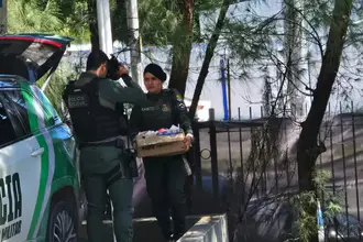 Dois policiais militares ao lado de uma viatura, sendo que uma agente carrega uma caixa de papelão