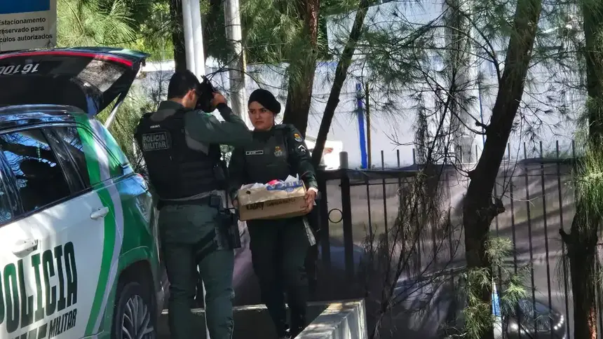 Dois policiais militares ao lado de uma viatura, sendo que uma agente carrega uma caixa de papelão