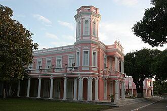 Prédio histórico da Reitoria da UFC, com fachada rosa e branca, torre central, colunas e jardins arborizados