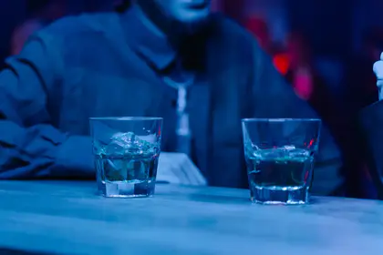 Dois copos com bebidas e gelo sobre uma mesa com uma pessoa ao fundo sob iluminaão azul